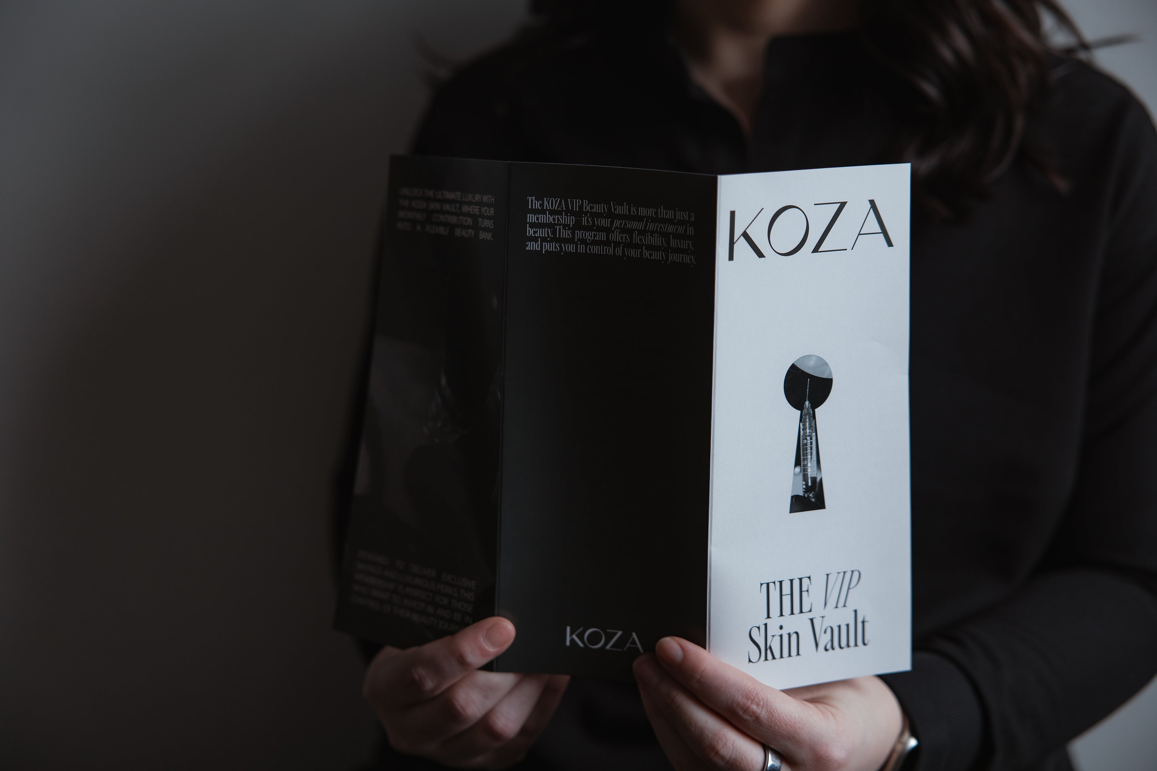 Person holding an open KOZA-branded 'The VIP Skin Vault' brochure at KOZA Skin Clinic, Port Coquitlam, Tri-Cities BC.